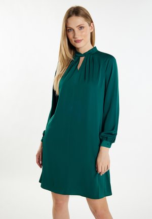 Femme aux longs cheveux blonds portant une robe vert foncé à manches longues avec un col haut et un détail en forme de serrure, debout sur fond blanc.
