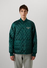 Chaqueta acolchada de color teal con botones a presión, cuello acanalado, dos bolsillos frontales y logotipo bordado en el lado izquierdo del pecho. Textura de satén suave.