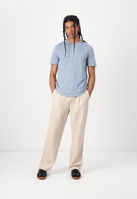 Camiseta de manga corta azul claro combinada con pantalones holgados beiges. El modelo lleva zapatos negros. Fondo minimalista.