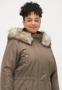 Parka castanha com acabamento de pêlo sintético na capucha, fecho frontal com zíper e cintura elástica. Apresenta dois bolsos no peito e detalhes em metal prateado.