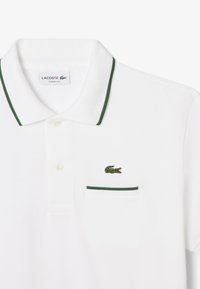 Polo bianco con accenti e colletto verdi. Presenta un piccolo logo di coccodrillo ricamato in verde e una tasca con bordatura verde. Vestibilità classica.