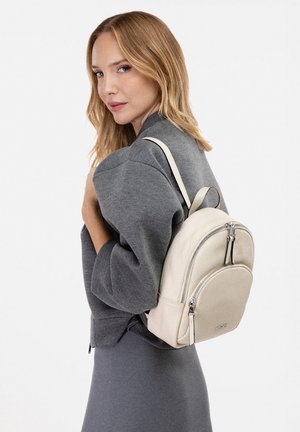 Femme aux cheveux blonds portant une tenue grise et un petit sac à dos en cuir blanc sur une épaule, sur fond blanc uni.