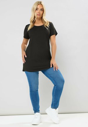 Frau mit blondem Haar, die eine schwarze Kurzarmtunika, blaue enge Jeans und weiße Sneakers trägt und vor einem einfachen hellen Hintergrund steht.