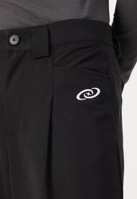 Černé kalhoty s detaily knoflíku a poutka na pásek, s malým bílým vyšitým logem na pravé přední kapse.