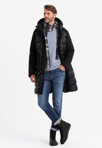 Schwarze Pufferjacke mit Kapuze, blauer Pullover, Jeans und schwarze Stiefel. Der Schal hat ein kariertes Muster in Blau, Grau und Rot.