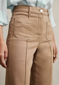 Pantaloni bej cu talie înaltă, cu buzunare în față și în spate, cu croială dreaptă și detalii de cusătură vizibile. Materialul pare robust și texturat.