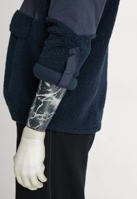 Marine fleece jack met textuurstof, contrasterende gladde schouder en arm, met een marmerpatroon prothese-arm en een witte handschoen.
