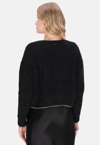 Vrouw met golvend blond haar, gekleed in een zwarte fuzzy trui en zwarte rok, van achteren afgebeeld tegen een effen lichte achtergrond.