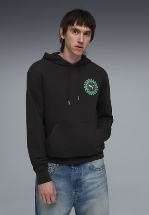 Jeune homme portant un sweat à capuche noir avec un logo turquoise en forme de soleil et un jean bleu délavé, debout devant un arrière-plan en dégradé de gris.