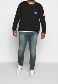 Sudadera negra de manga larga con el logo de NASA y un parche de la bandera americana, combinada con unos vaqueros claros desgastados y zapatillas blancas.