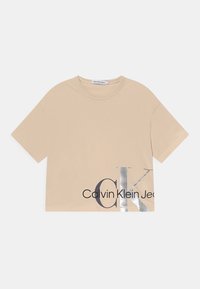 Camiseta corta de manga corta color beige con cuello redondo que presenta una gran impresión en plata y negro de "CK" y "Calvin Klein Jeans" en la parte inferior derecha del frente.