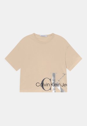 Krátký béžový crop top s krátkým rukávem a kulatým výstřihem, na přední dolní pravé straně s velkým stříbrným a černým potiskem "CK" a "Calvin Klein Jeans".