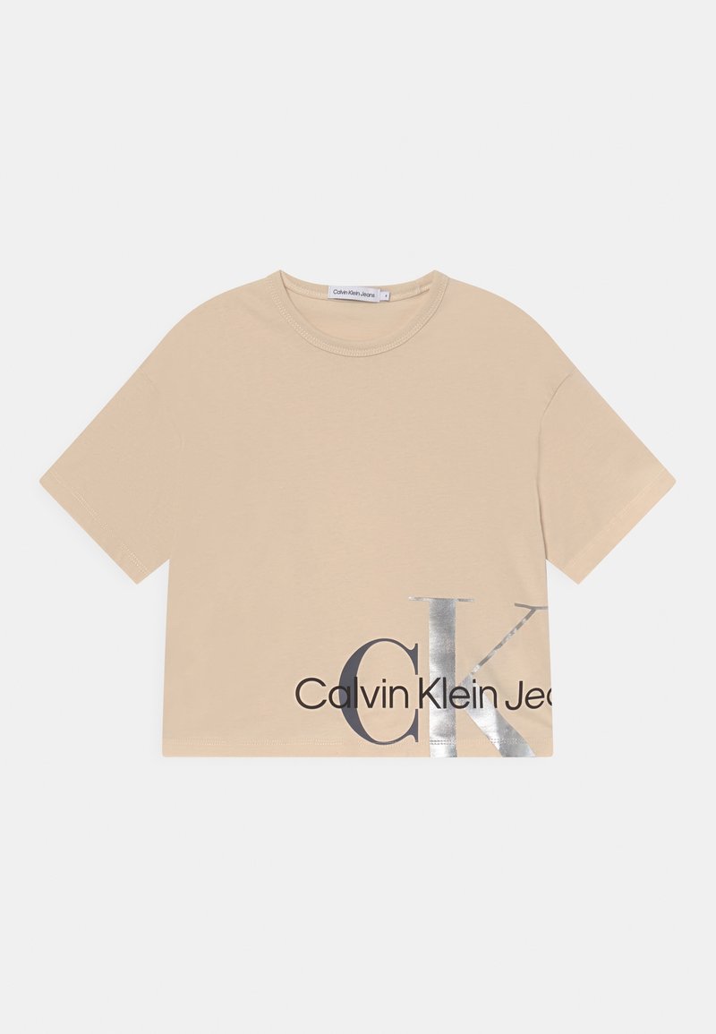 Camiseta corta de manga corta color beige con cuello redondo que presenta una gran impresión en plata y negro de "CK" y "Calvin Klein Jeans" en la parte inferior derecha del frente.