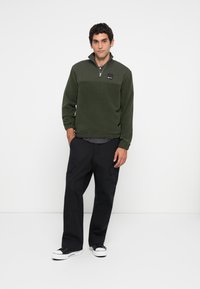 Zöld fleece pulóver, fél cipzárral, fekete logóval ellátott folttal. Fekete cargo nadrággal és fekete-fehér sportcipővel párosítva.