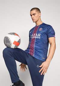 Maillot de football bleu avec des motifs géométriques rouges, arborant le logo de Qatar Airways. Modèle portant un pantalon de sport bleu marine, tenant un ballon à motifs.