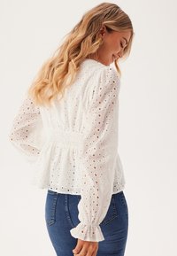 Blusa bianca con tagli a occhiello, vita arricciata e maniche a palloncino. Indossata con jeans blu aderenti, tessuto testurizzato e polsini delle maniche volant.