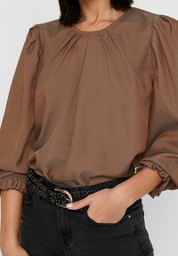 Brązowa bluzka z długim rękawem, z marszczonymi szwami na ramionach i okrągłym dekoltem. Czarny pasek z srebrnymi ćwiekami noszony na czarnych dżinsach.