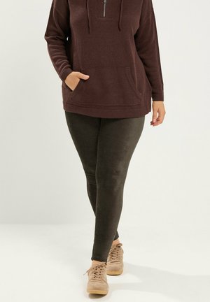 Person iført en brun hættetrøje, mørke leggings og beige sneakers stående mod en ensfarvet lys baggrund.