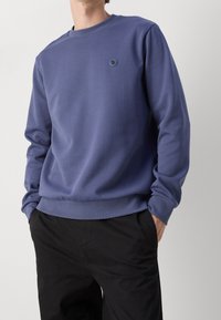 Blå sweatshirt med rund halsringning, texturerad tyg, ribbade manchetter och nederkant, som har en liten blå logotyp på bröstet. Bärs med svarta byxor.