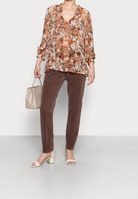 Blusa de estampado floral en tonos cálidos con un dobladillo con volantes, combinada con pantalones marrones texturizados y sandalias blancas de tacón cuadrado. Accesorio: bolso beige.