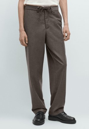 Persona che indossa pantaloni larghi marroni con cordoncino e scarpe eleganti in pelle nera, in piedi contro uno sfondo chiaro e uniforme.