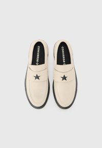 Beige slip-on sko med en svart stjerneaccent på stroppen, med en teksturert gummisåle og svart indre fôr.