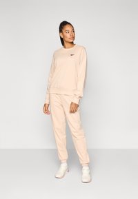 Bluză bej și pantaloni sport asortați, confecționați dintr-un material moale. Bluza are guler rotund; pantalonii sport au manșete elastice. Pantofi sport albi.