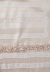 Écharpe beige clair avec un tissage diagonal, ornée de rayures blanches et de bords à franges. Le nom de la marque "MICHAEL KORS" est subtilement tissé dans le design.