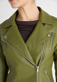 Personne portant une veste en cuir vert olive avec des fermetures éclair et des pressions argentées sur un haut brun foncé, montrant le cou et le bas du visage.