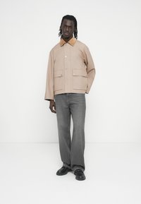 Veste beige avec un col marron texturé, dotée de deux poches avant et de boutons-pression argentés, associée à un jean gris à jambe large et des chaussures noires.