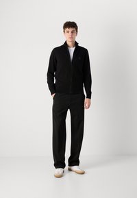 Svart zip-jacka med hög krage, tillsammans med lösa svarta byxor och vita sneakers. Slät tyg, minimal branding på jackan.