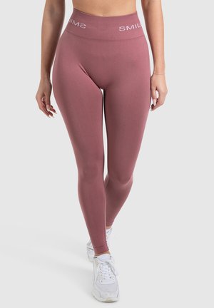 Hoge roze leggings met een textuurafwerking en "SMIL" branding op de tailleband, gecombineerd met witte sportschoenen.