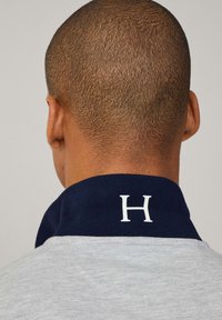 Šedé polokošile s tmavě modrým límečkem, na zadní straně s bílým písmenem "H". Tkání vypadá hladce a ležérně.