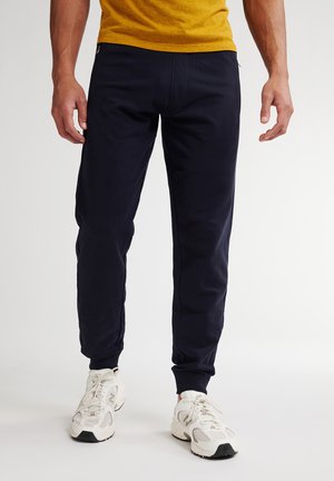 Hombre con pantalones jogger negros, zapatillas blancas y camiseta mostaza, de pie sobre un fondo blanco liso.