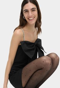 Černé šaty s velkou mašlí, se zdobenými ramínky, kombinované s černými punčochovými legíny s tečkami. Hladká tkanina a přiléhavý střih.