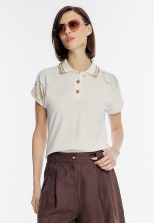 Kobieta ubrana w kościelną koszulkę polo z kwiatowym haftem i złotymi guzikami, połączoną z brązowymi spodniami z wysokim stanem i okularami przeciwsłonecznymi.