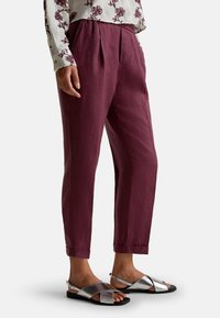 Personne portant un pantalon court plissé violet taille haute avec des ourlets retroussés et des sandales métalliques argentées à bout ouvert, avec un haut blanc à motifs.