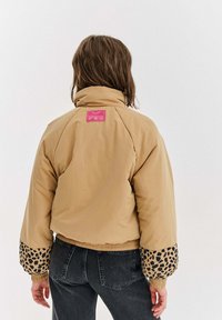 Osoba s vlasmi po pleciach, hnedými, oblečená v béžovej bunde s leopardím vzorom na manžetách a ružovou etiketou na hornej časti chrbta, doplnená tmavými džínami.