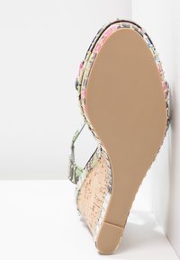 Sandale à talon compensé en liège, brides multicolores, clous dorés, et semelle lisse beige, présentée en angle contre un fond blanc en coin.