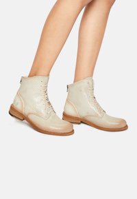 Botas de tobillo de color beige con perforaciones, cordones en la parte delantera, suela de madera y detalles lisos en el talón. Presenta un cierre lateral para ponerlas y quitarlas con facilidad.