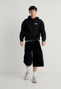 Νέος άνδρας που περπατά φορώντας μαύρο φούτερ με κουκούλα, φαρδιά μαύρη βερμούδα, λευκές κάλτσες και λαμπερά ασημί αθλητικά παπούτσια, μπροστά σε απλό λευκό φόντο.