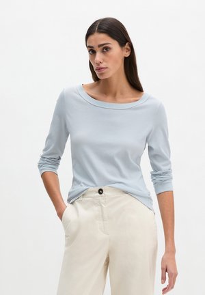 Vrouw met lang donker haar, gekleed in een lichtblauw shirt met lange mouwen en beige hoge taillebroek, staand met één hand in haar zak.