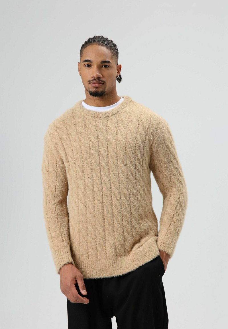 Beige kabelgebreide trui gemaakt van zacht, pluizig materiaal met lange mouwen en een ronde hals, gecombineerd met een zwarte broek.