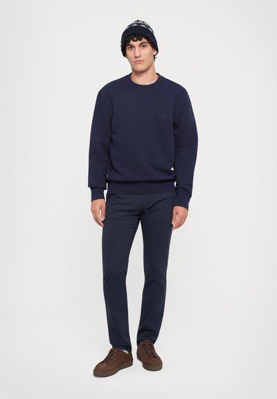 Felpa girocollo navy e pantaloni abbinati, completati da sneakers marroni. Un berretto di lana lavorato a maglia con motivo completa il look. Il materiale ha una finitura testurizzata.