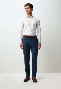 Camicia bianca a maniche lunghe con colletto button-down, abbinata a pantaloni su misura blu scuro e scarpe di pelle marrone, con cintura marrone.