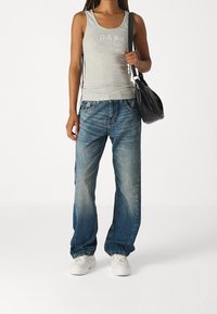Camiseta sin mangas gris con el logo "GAP", combinada con unos vaqueros de mezclilla azules de corte holgado. El modelo lleva un bolso negro de hombro y calza zapatillas blancas.