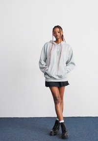 Jeune femme avec de longues tresses portant un sweat à capuche gris surdimensionné, une jupe noire, des chaussettes blanches et des bottes noires, debout sur un tapis bleu devant un mur blanc.
