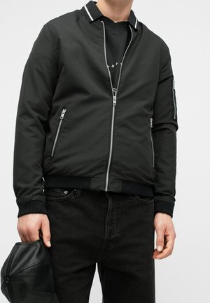 Personne portant une veste zippée noire par-dessus une chemise noire et un jean noir, tenant une casquette en cuir noire dans sa main gauche.