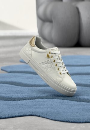 Biały skórzany sneaker ze złotymi akcentami i tłoczonym wzorem, umieszczony na teksturowanym niebieskim dywanie, na tle szarego mebla z poduszkami.