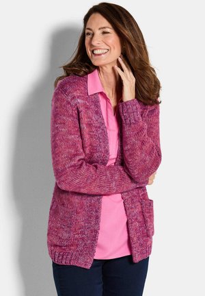Femme souriante touchant ses cheveux, portant une chemise rose à col et un cardigan en tricot violet avec des poches.